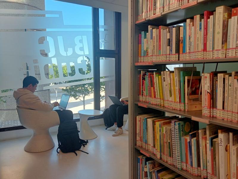 Un joven utiliza un portátil delante de una cristalera en la que se lee BJC Cubit. En primer plano hay una estantería con libros.